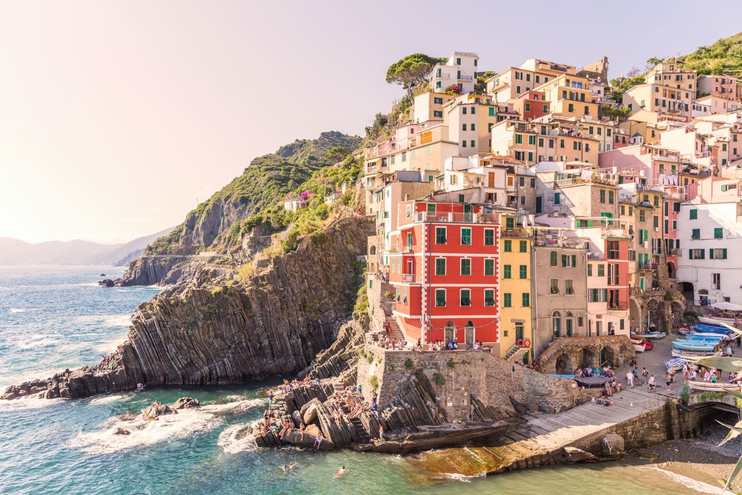 Colorful buildings on a cliff overlooking the sea in Riomaggiore, Cinque Terre, with people enjoying the scenic view.
