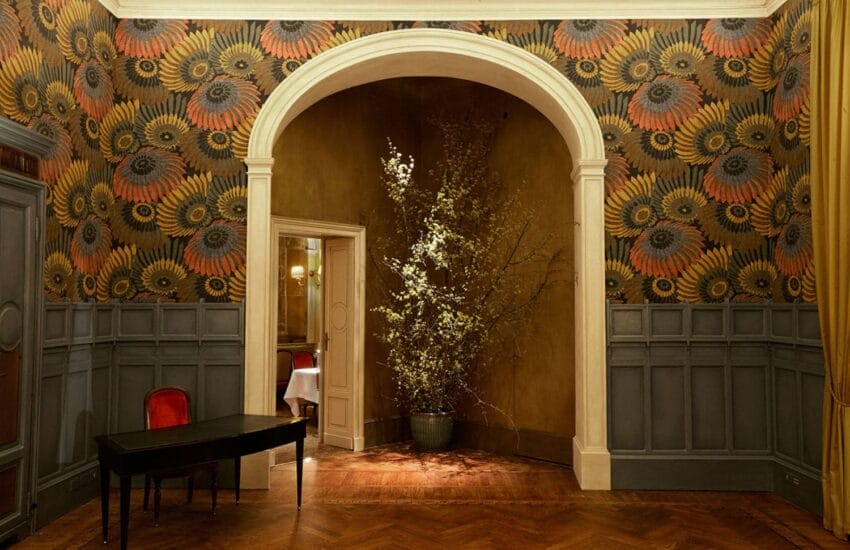 Elegant vintage room with patterned walls, arched doorway, potted plant, dark table, red chair, and chevron wood floor.