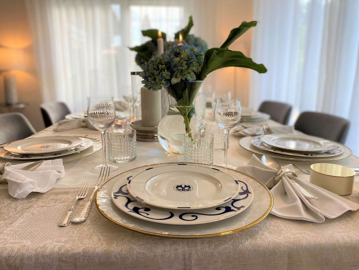 Elegant dining table set with white and blue patterned plates, crystal glassware, and a floral centerpiece.