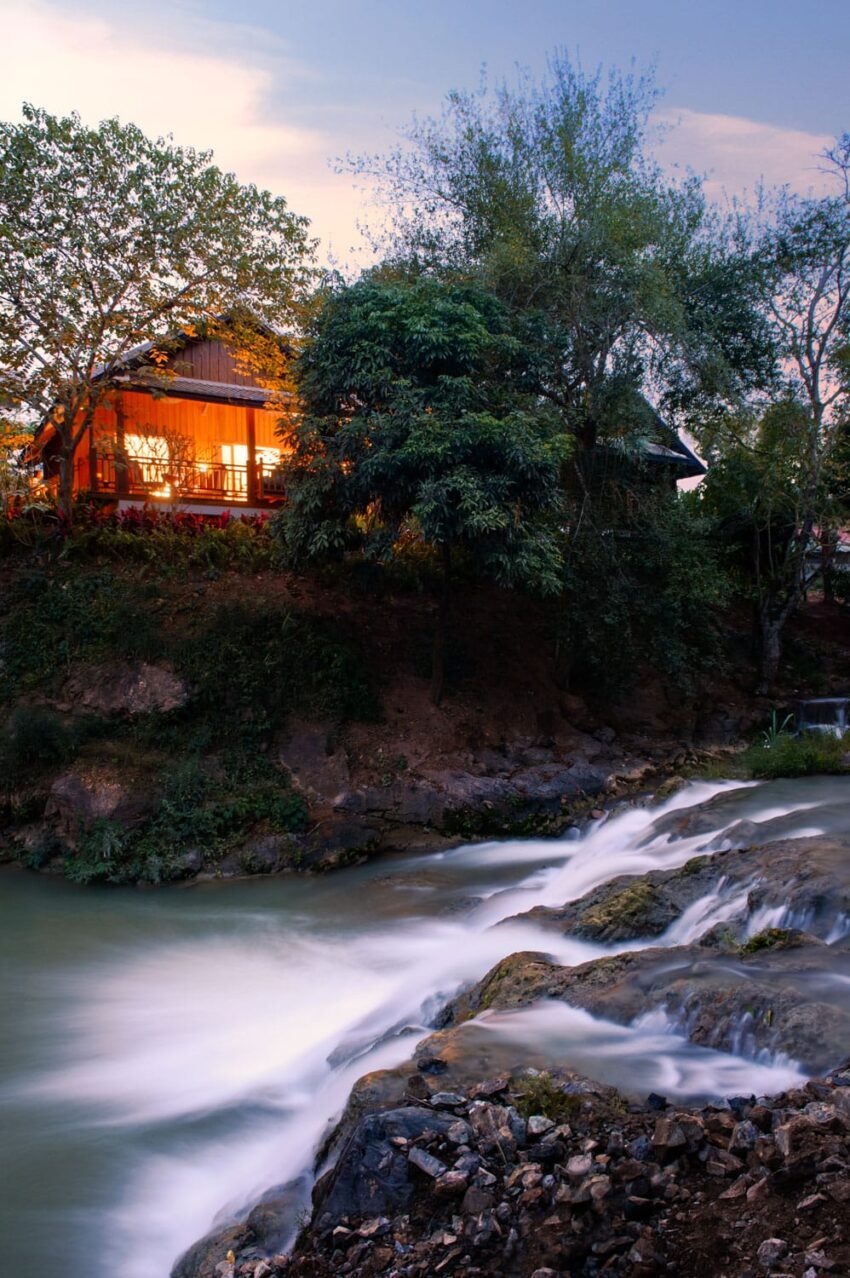 House with warm lights by a river surrounded by trees at dusk.