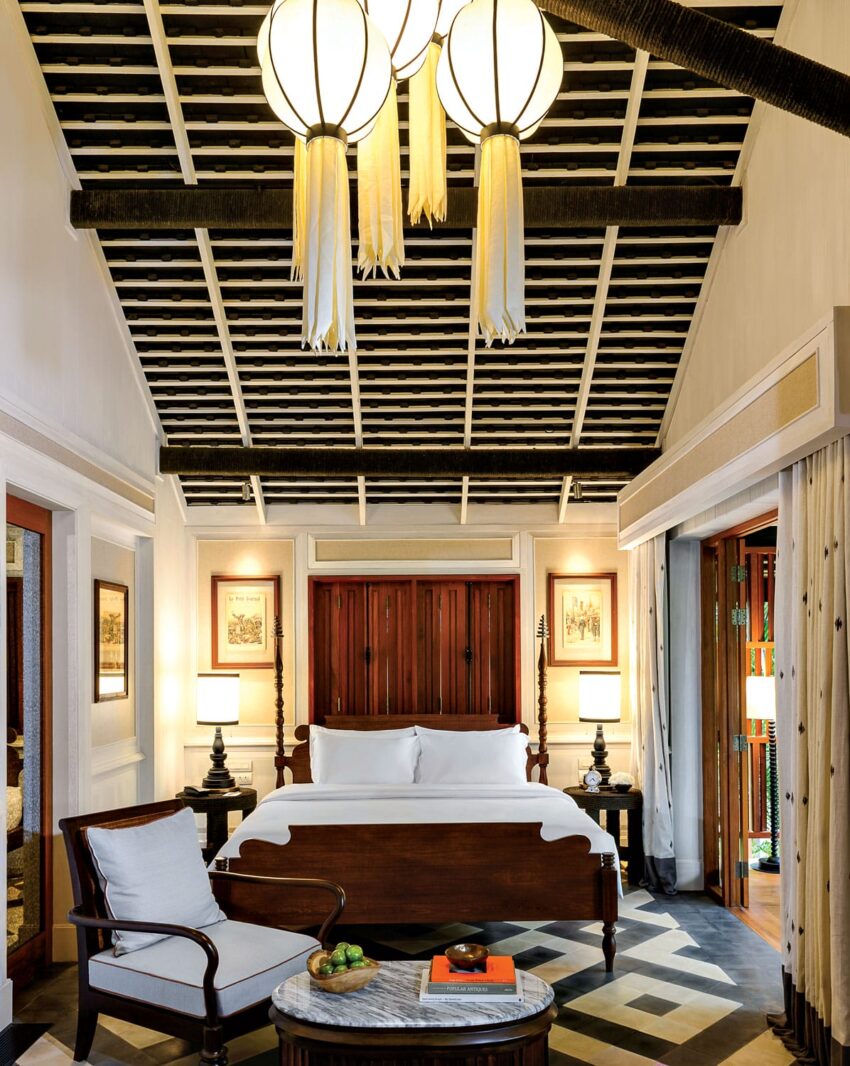 Luxurious bedroom with a wooden canopy bed, elegant decor, armchair, and a round marble table under large hanging lights.