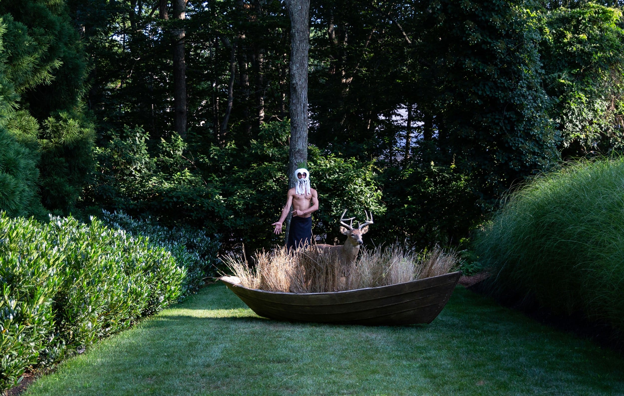 Person with a mask stands beside a wooden boat planter with grasses and a deer statue in a lush garden setting.