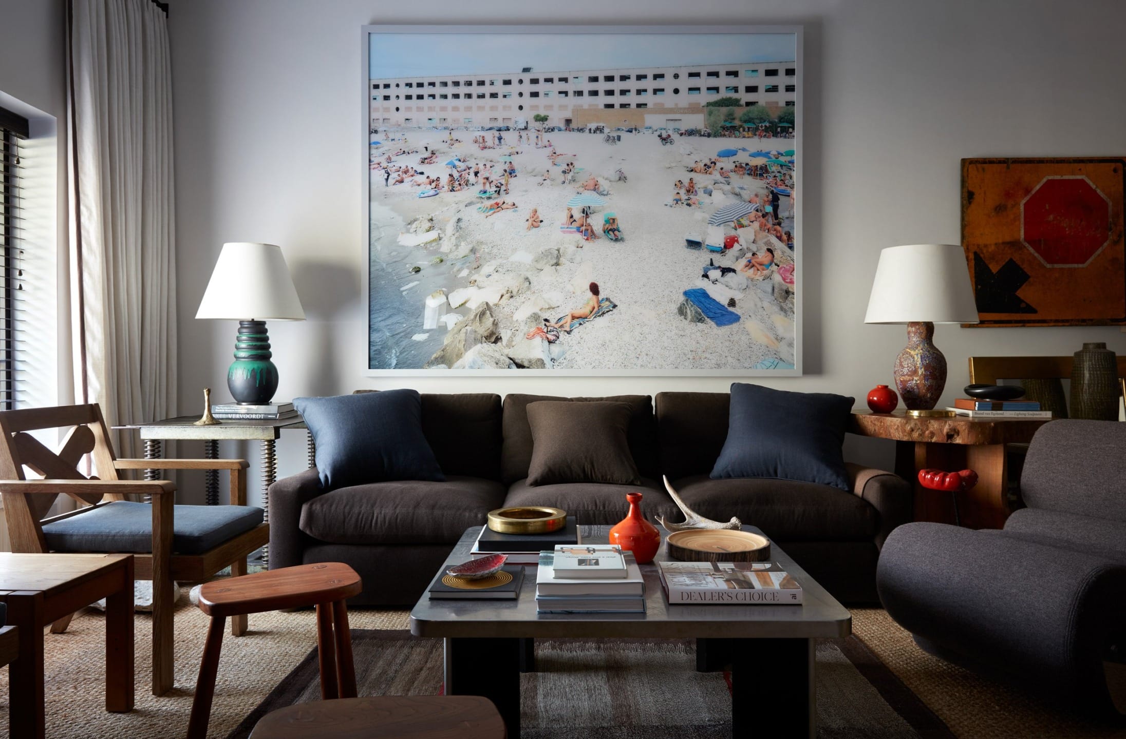 Cozy living room with dark sofa, large beach photo on wall, various chairs, tables, lamps, and decorative objects.