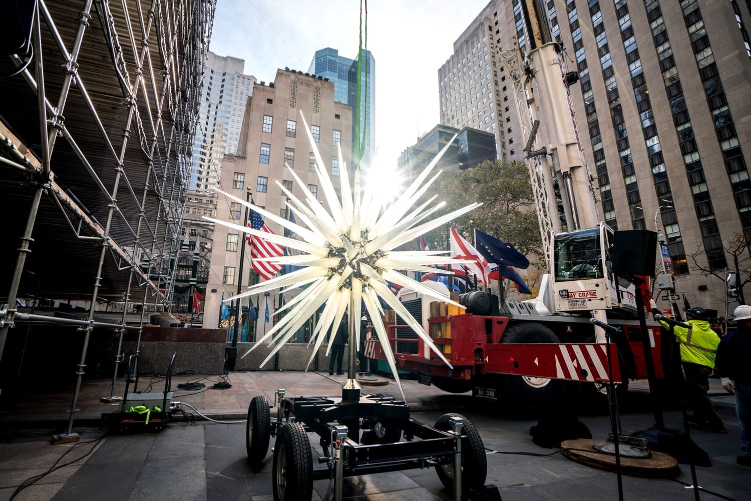 Large star sculpture being installed at Rockefeller Center with cranes and American flags in the background.