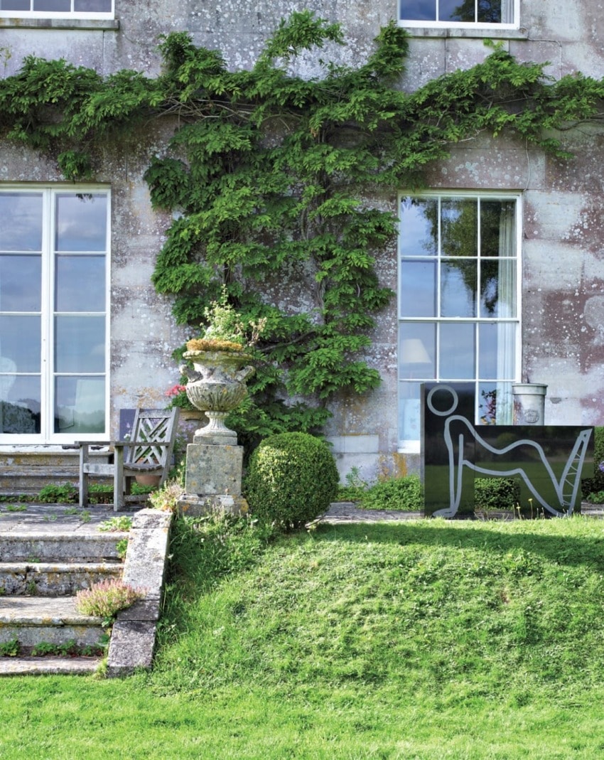 Garden with stone steps, a sculpted hedge, outdoor seating, and a wall covered in green vines near large windows.