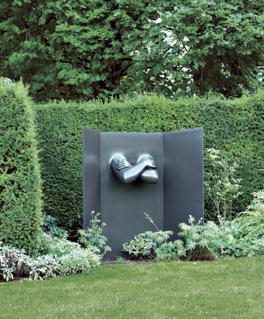 Contemporary sculpture of a face and hands emerging from a metal screen in a lush garden setting.