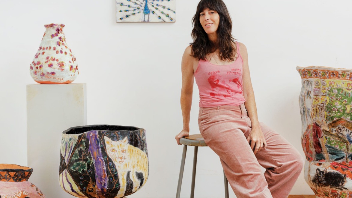 Person sitting on a stool surrounded by colorful, artistic pottery pieces in a studio setting.