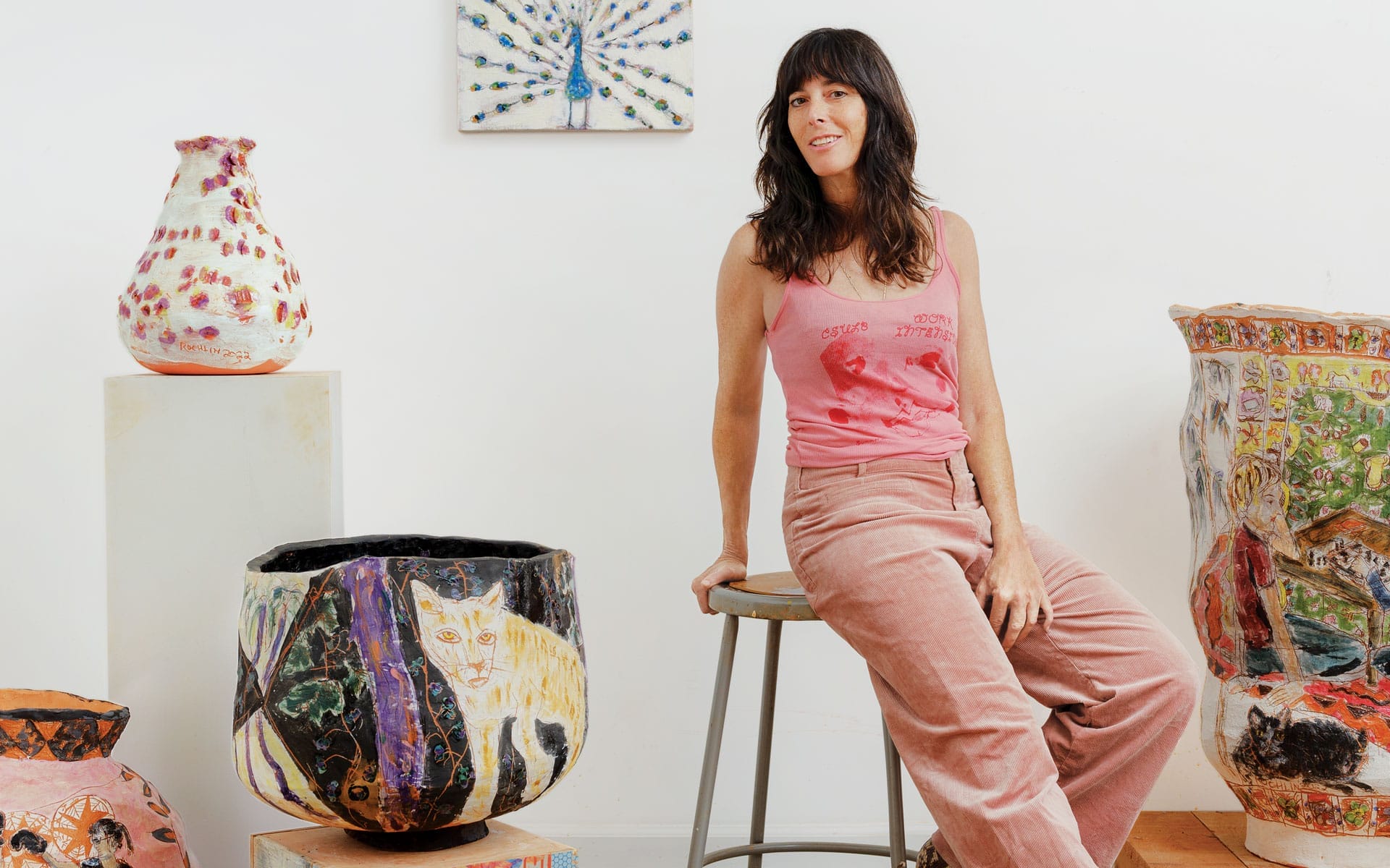 Person sitting on a stool surrounded by colorful, artistic pottery pieces in a studio setting.