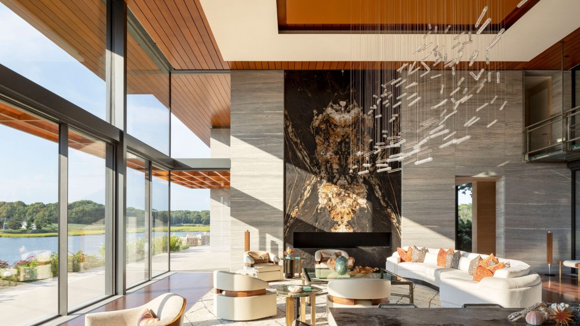 Modern living room with large windows, abstract chandelier, elegant furnishings, and a view of a lake and greenery outside.