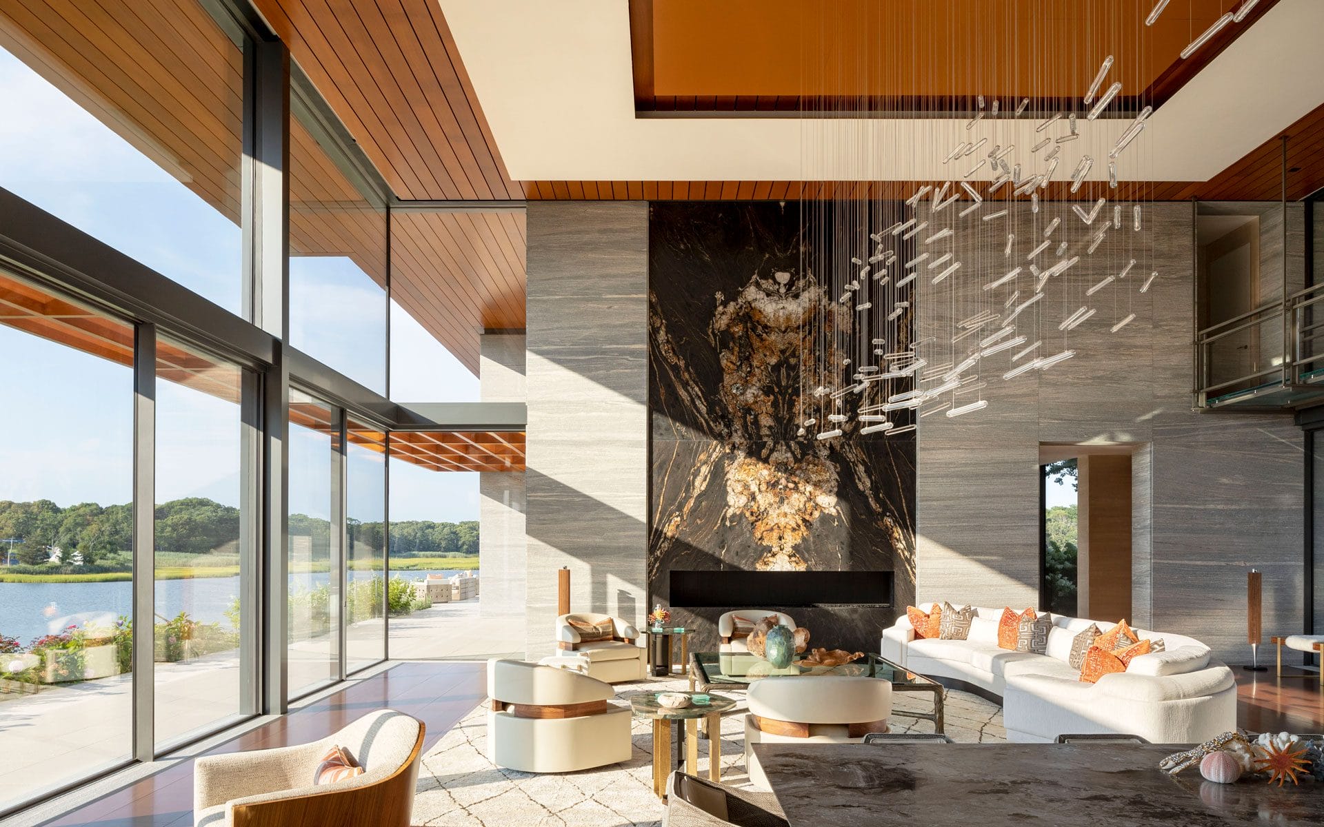 Modern living room with large windows, abstract chandelier, elegant furnishings, and a view of a lake and greenery outside.