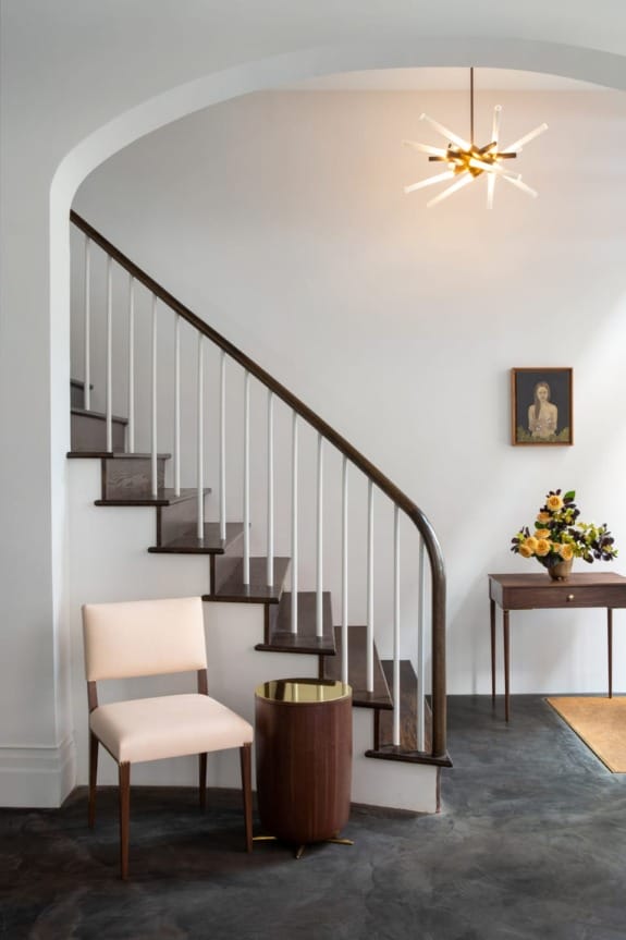 Modern interior with curved staircase, elegant chair, small round table, pendant light, and a floral arrangement on a console table.