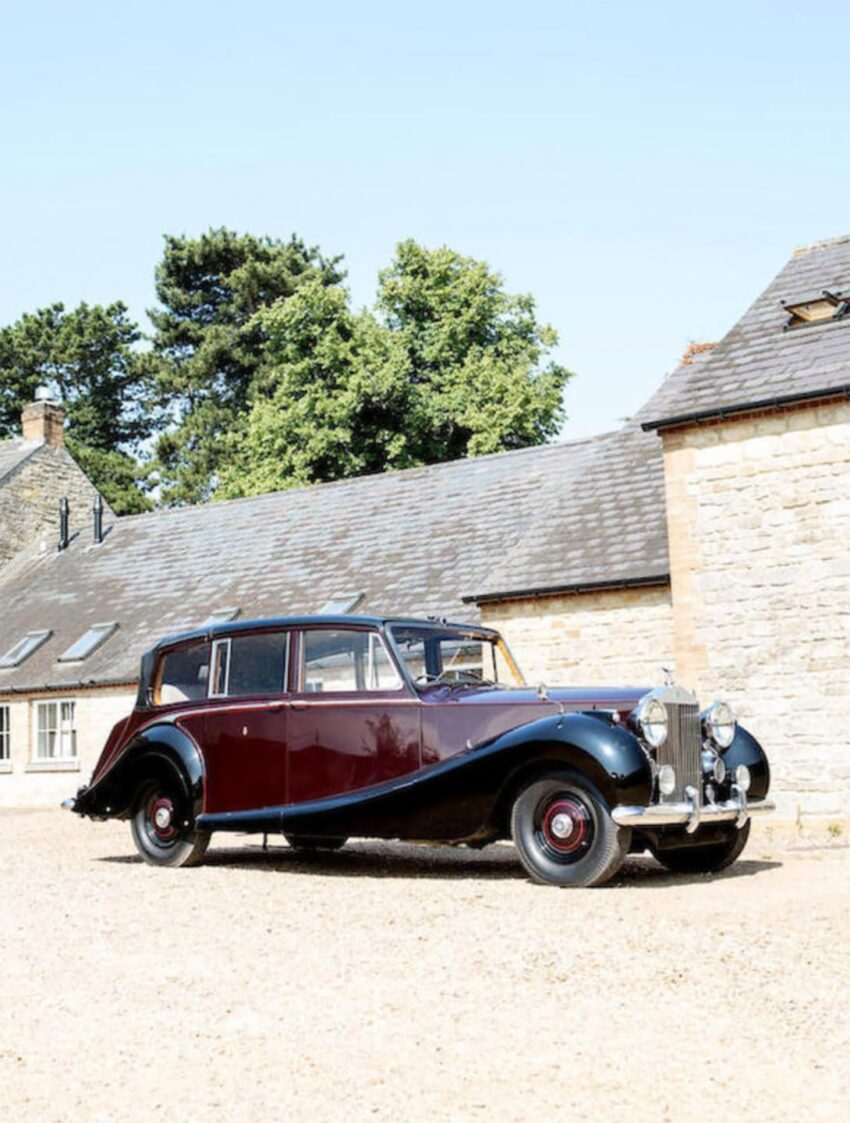 Vintage black and maroon luxury car parked in front of a stone building with a clear blue sky.