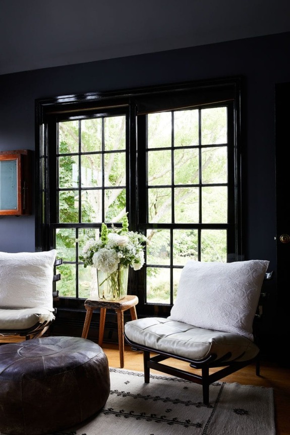 Modern living room with large windows, white chairs, a brown ottoman, and a small table with a floral arrangement.