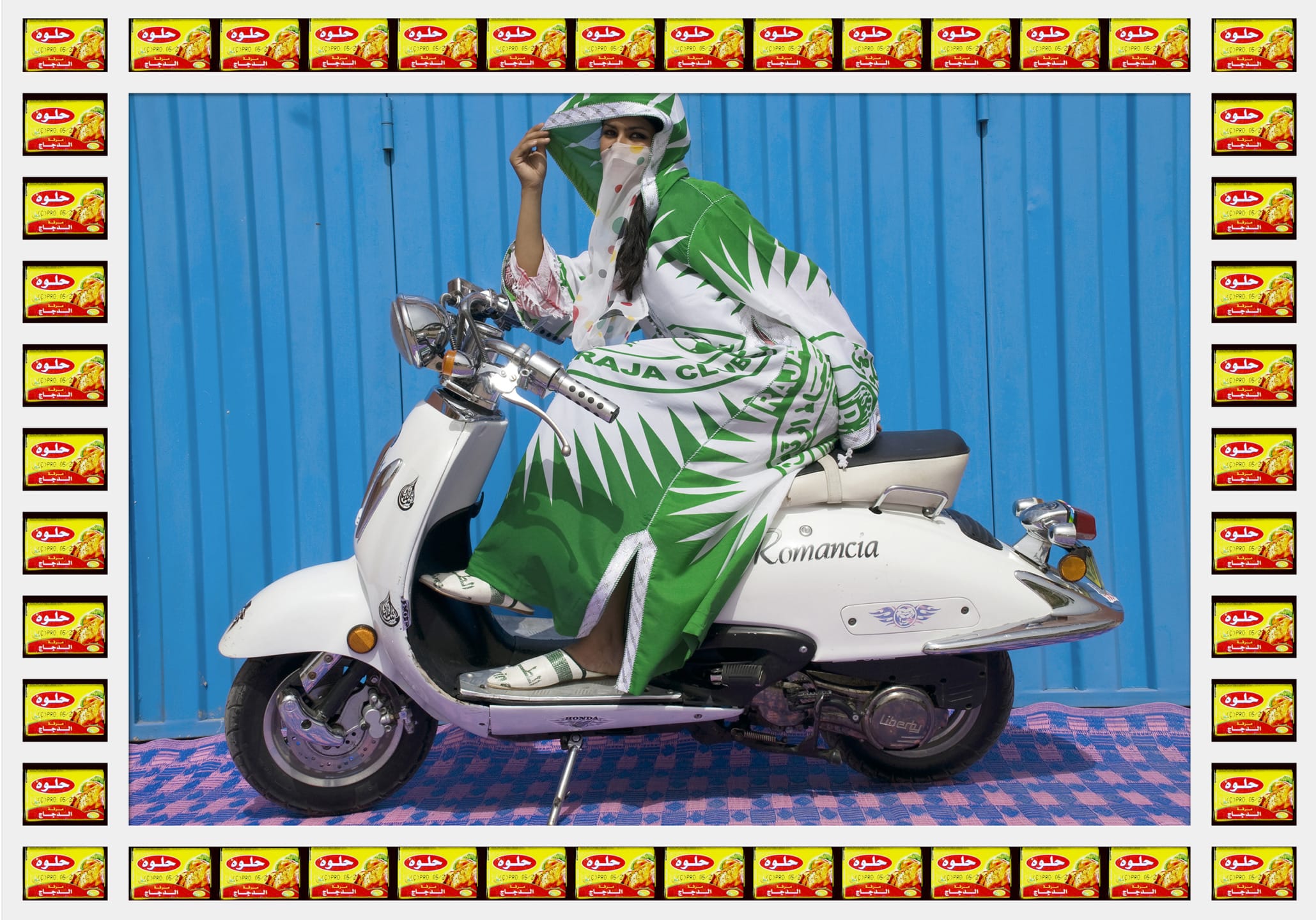 Person draped in green and white fabric poses on a white scooter against a blue background, framed by vibrant patterns.