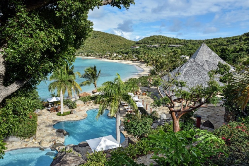 Tropical beach resort with palm trees, turquoise pool, and lush greenery overlooking a serene ocean bay and distant hills.