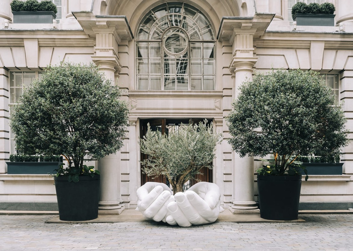 Large white sculpture of hands in a courtyard flanked by two large potted trees in front of a grand building entrance.
