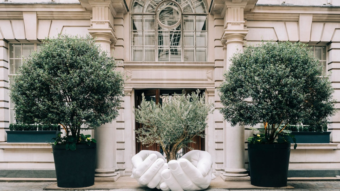 Large sculpture of interlocking hands displayed between two potted trees at a building entrance with classical architecture.