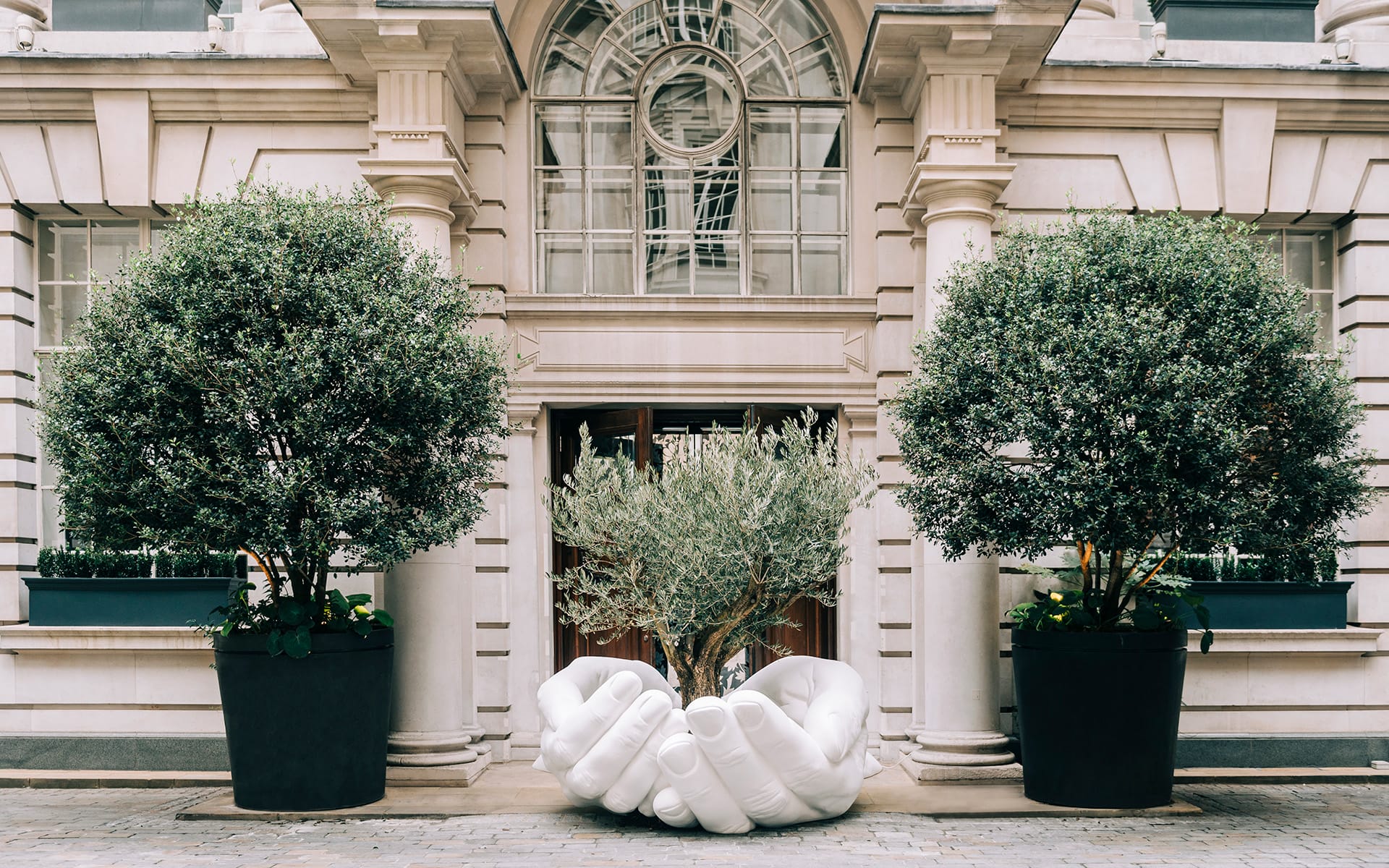 Large sculpture of interlocking hands displayed between two potted trees at a building entrance with classical architecture.