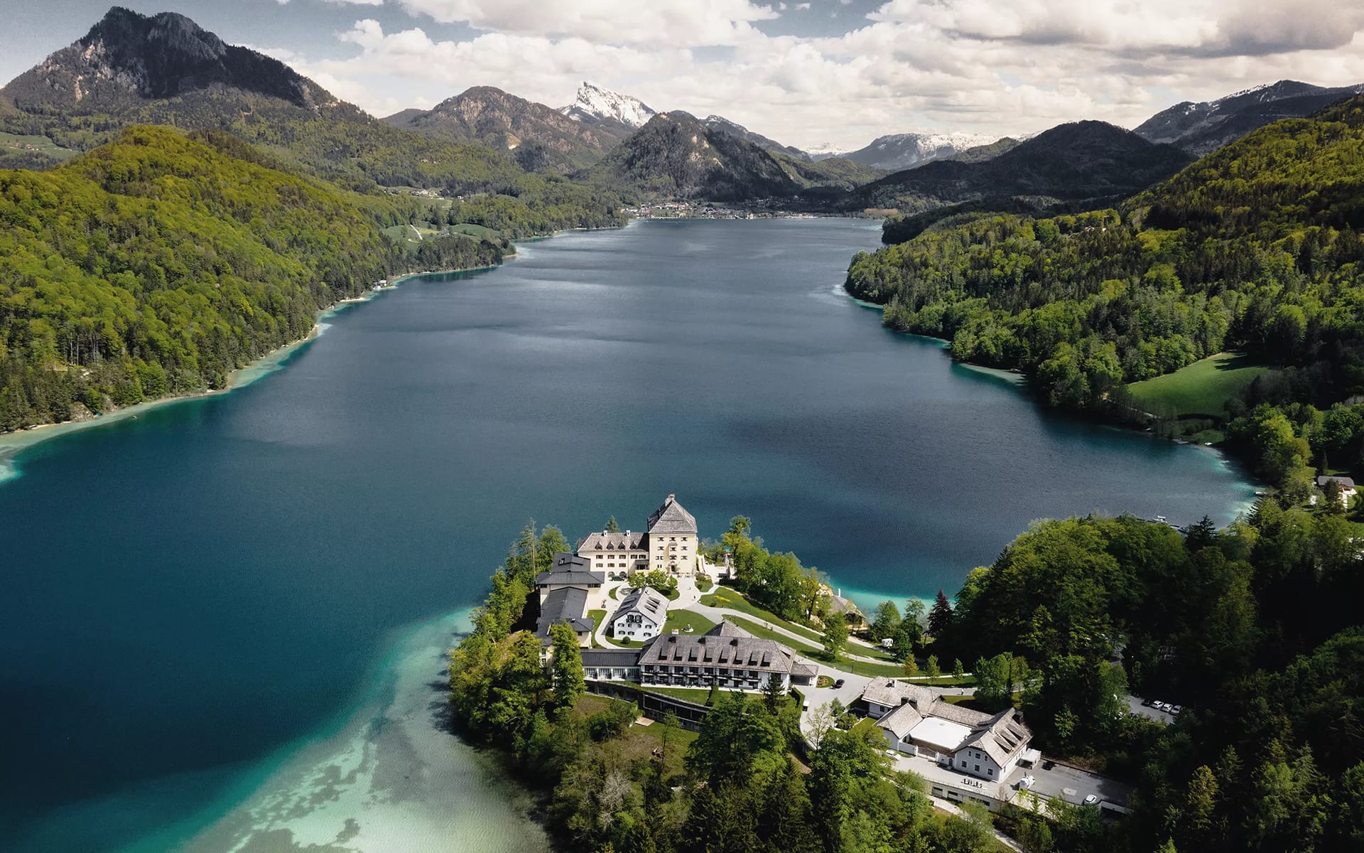 Aerial view of an alpine lake surrounded by green forests and mountains with a castle-like building near the shore.