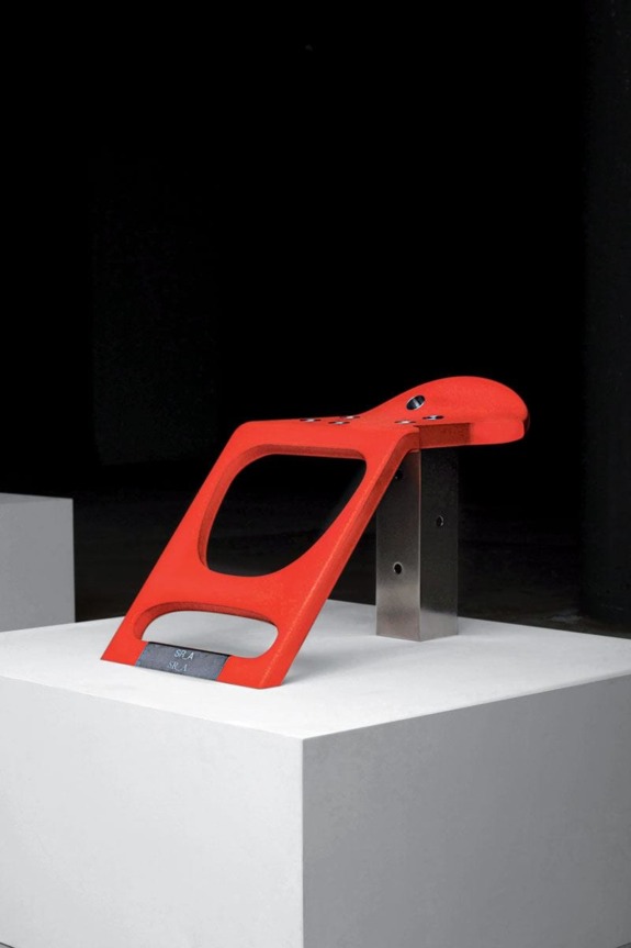 Modern red abstract sculpture on a white pedestal against a dark background in an art gallery setting.