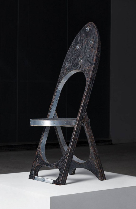 Iron chair sculpture on display, featuring a unique, industrial design with circular cutouts, on a white pedestal.