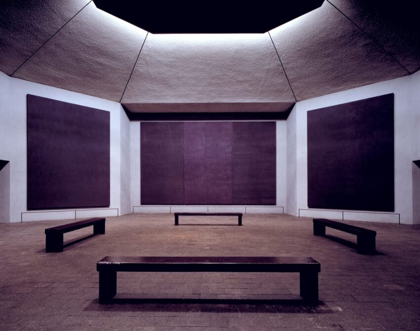 Interior of a modern art space with large dark paintings on walls and wooden benches on a stone floor.