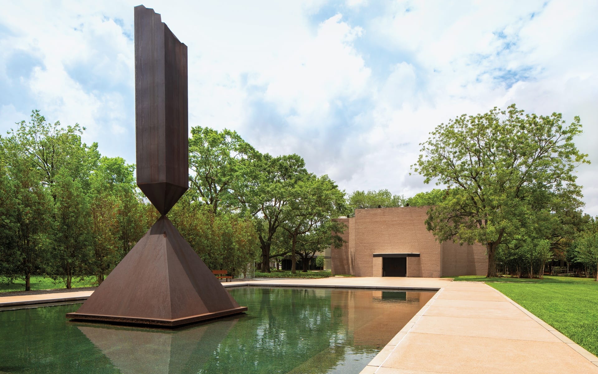 Weathered steel sculpture reflecting in a tranquil pool with trees and a modern brick building in the background on a cloudy day.