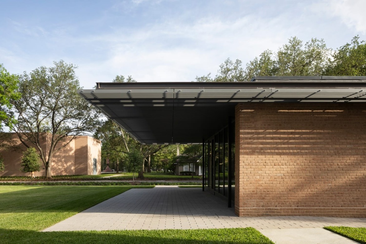 Modern building with brick facade, overhanging roof, and glass walls, surrounded by trees and a grassy landscape on a sunny day.