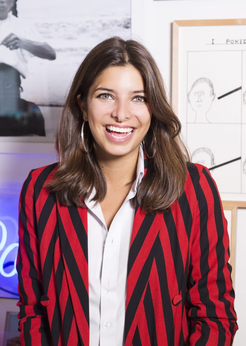 Person smiling, wearing a red and black striped jacket over a white shirt, with framed pictures in the background.
