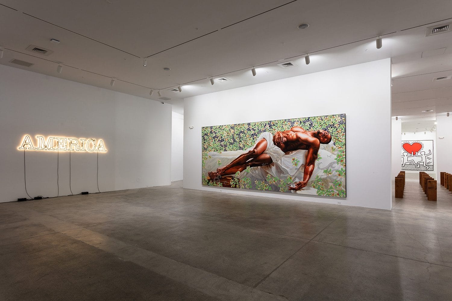 Art gallery interior with illuminated "America" sign, large painting of a reclining figure, and small graphic with a heart and figure.