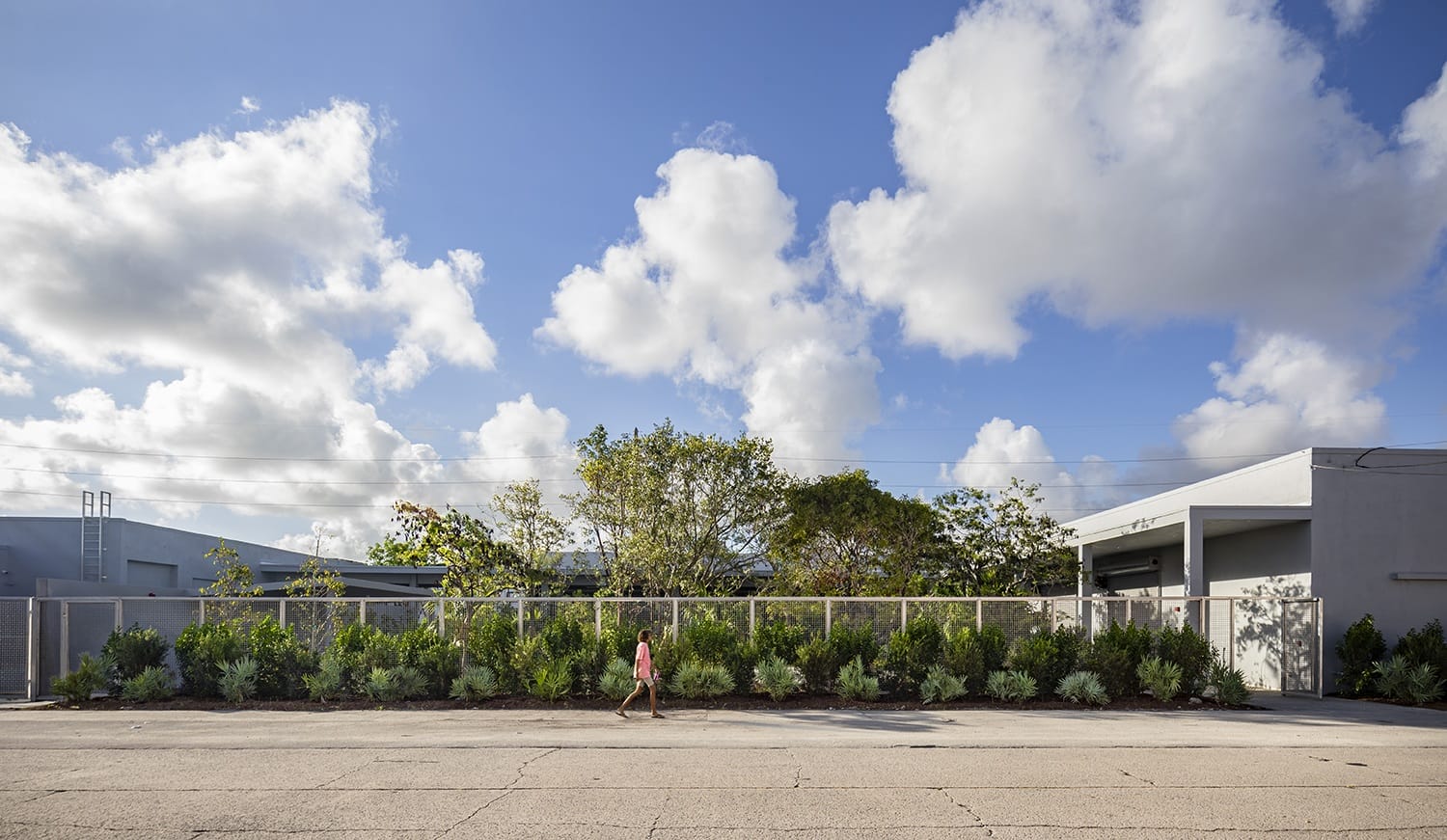 Person walking on street beside fenced area with greenery, buildings, and cloudy sky in the background.