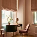 Cozy room with oval desk, vase with flowers, lamp, and pink chair, two windows with beige curtains, geometric rug on floor