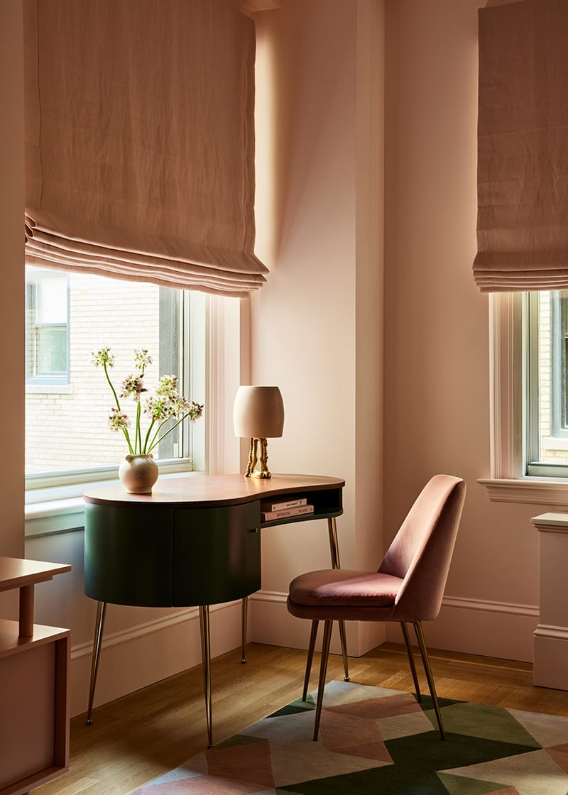 Cozy room with oval desk, vase with flowers, lamp, and pink chair, two windows with beige curtains, geometric rug on floor