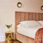 Plush pink bed with soft pillows, gold side table, pink tulips in vase, and geometric rug in cozy bedroom setting.