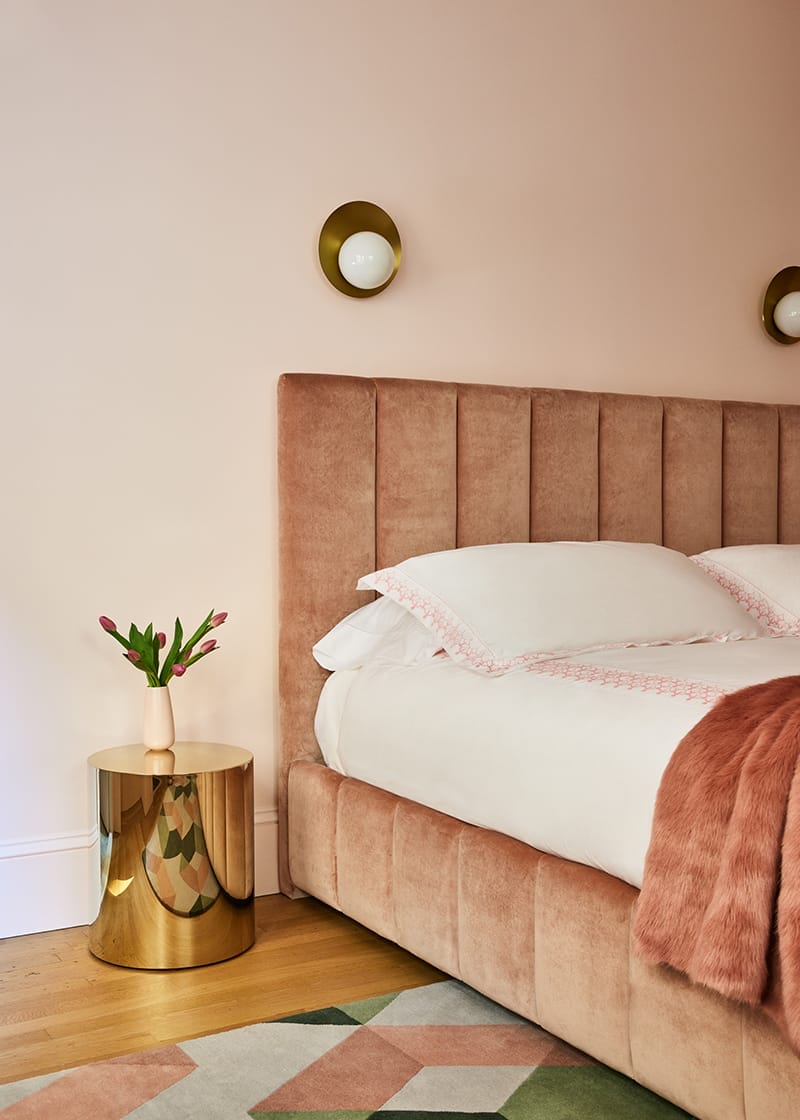 Plush pink bed with soft pillows, gold side table, pink tulips in vase, and geometric rug in cozy bedroom setting.