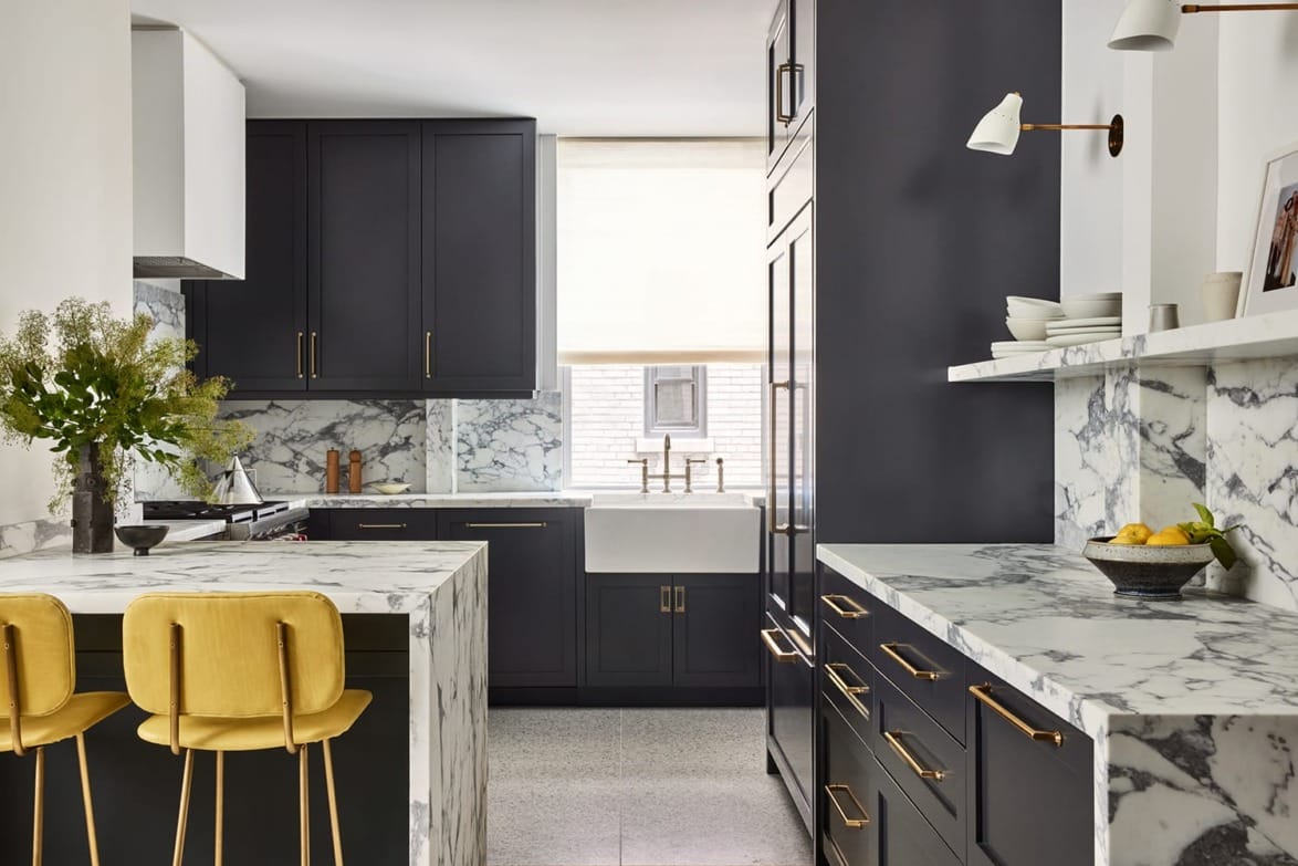 Modern kitchen with dark cabinets, marble countertops, and yellow bar stools, featuring a farmhouse sink and bright window.