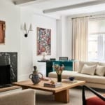Modern living room with a black marble fireplace, beige sofas, a wooden coffee table, and colorful artwork on white walls.
