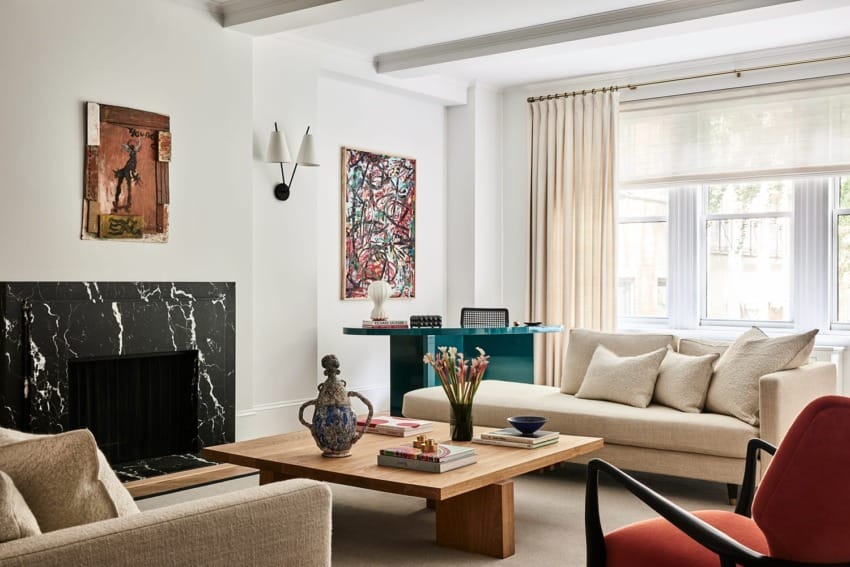 Modern living room with a black marble fireplace, beige sofas, a wooden coffee table, and colorful artwork on white walls.