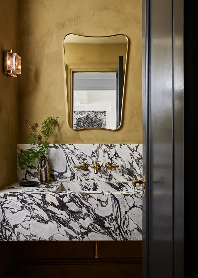 Modern bathroom with marble sink, gold accents, ornate mirror, and decorative vase with green plant.