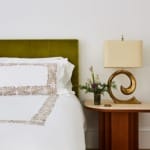 Bedroom with green headboard, ornate white bedding, unique gold lamp, and a wooden side table with a small plant arrangement.