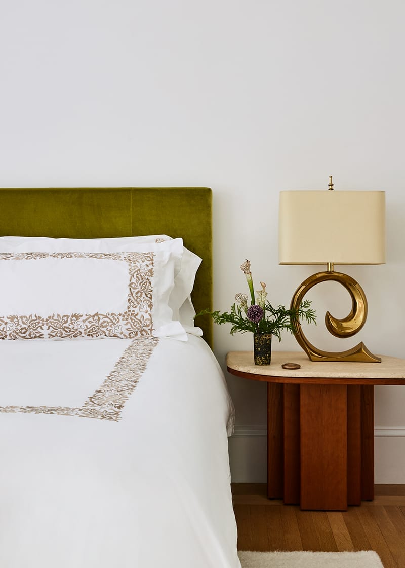 Bedroom with green headboard, ornate white bedding, unique gold lamp, and a wooden side table with a small plant arrangement.