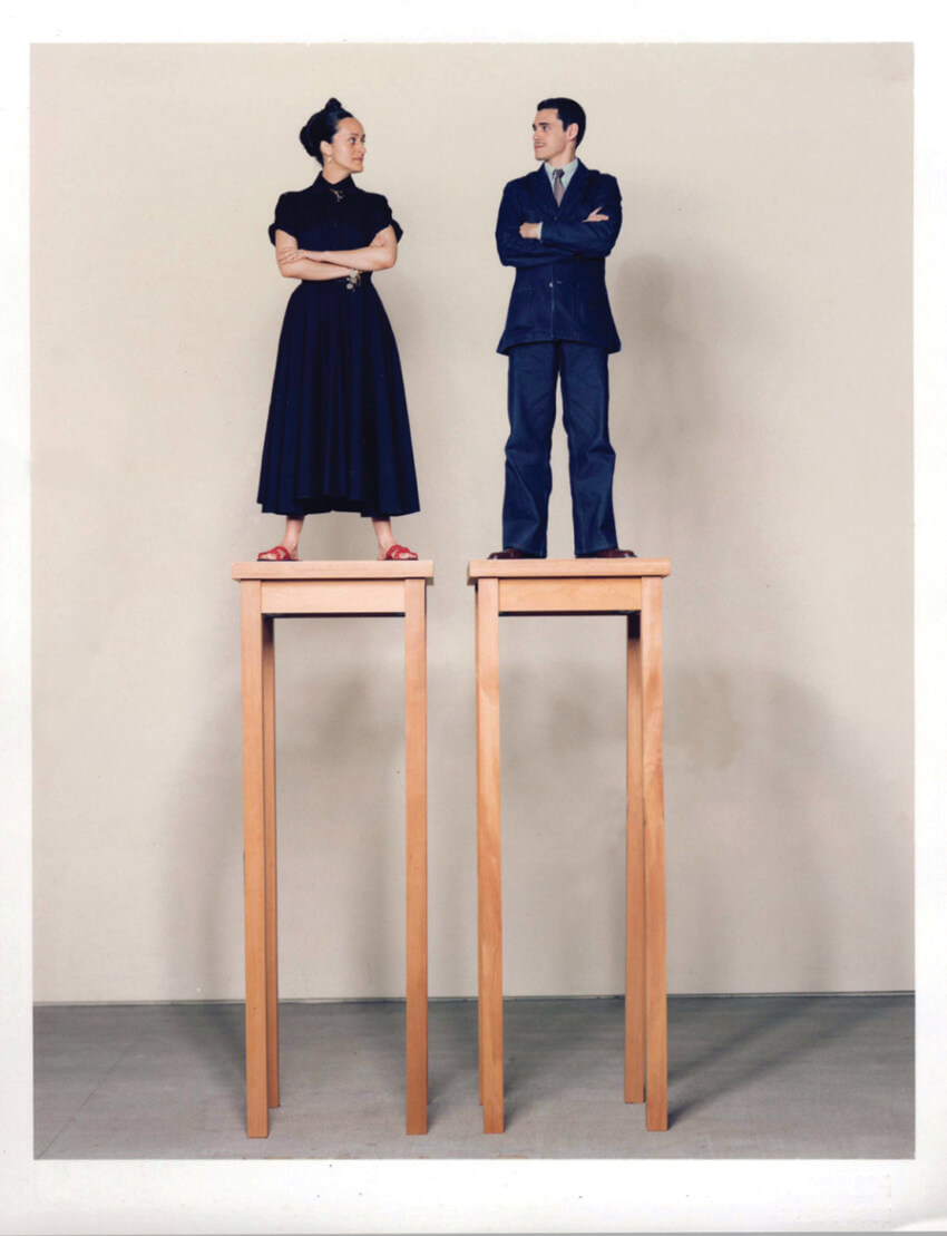 Man and woman in formal attire stand on tall wooden platforms against a plain backdrop, facing each other with crossed arms.