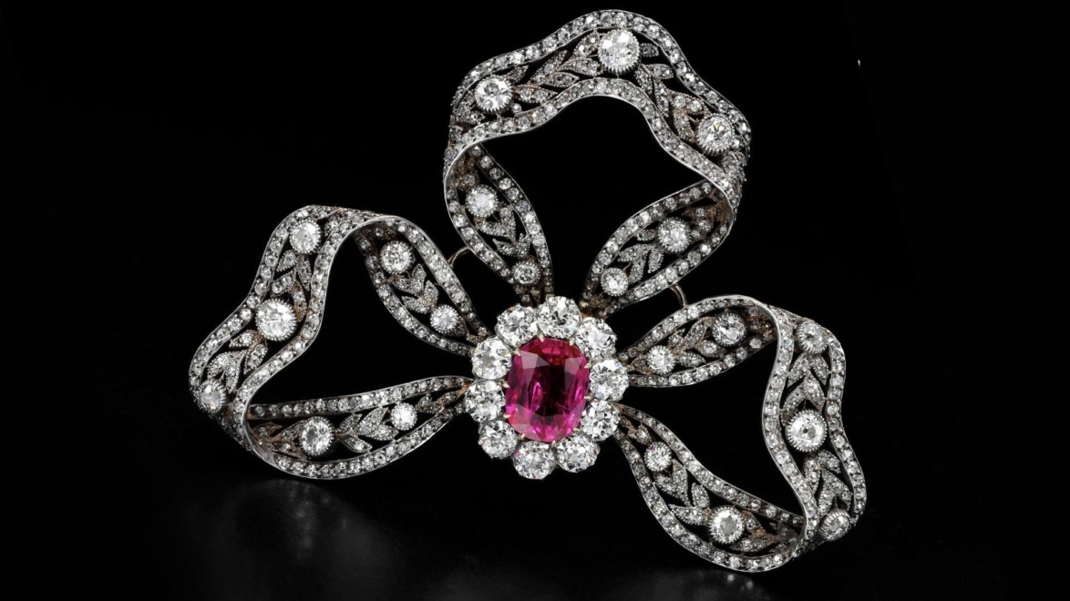 Elegant bow-shaped brooch with sparkling diamonds and a central vivid pink gemstone on a black background.