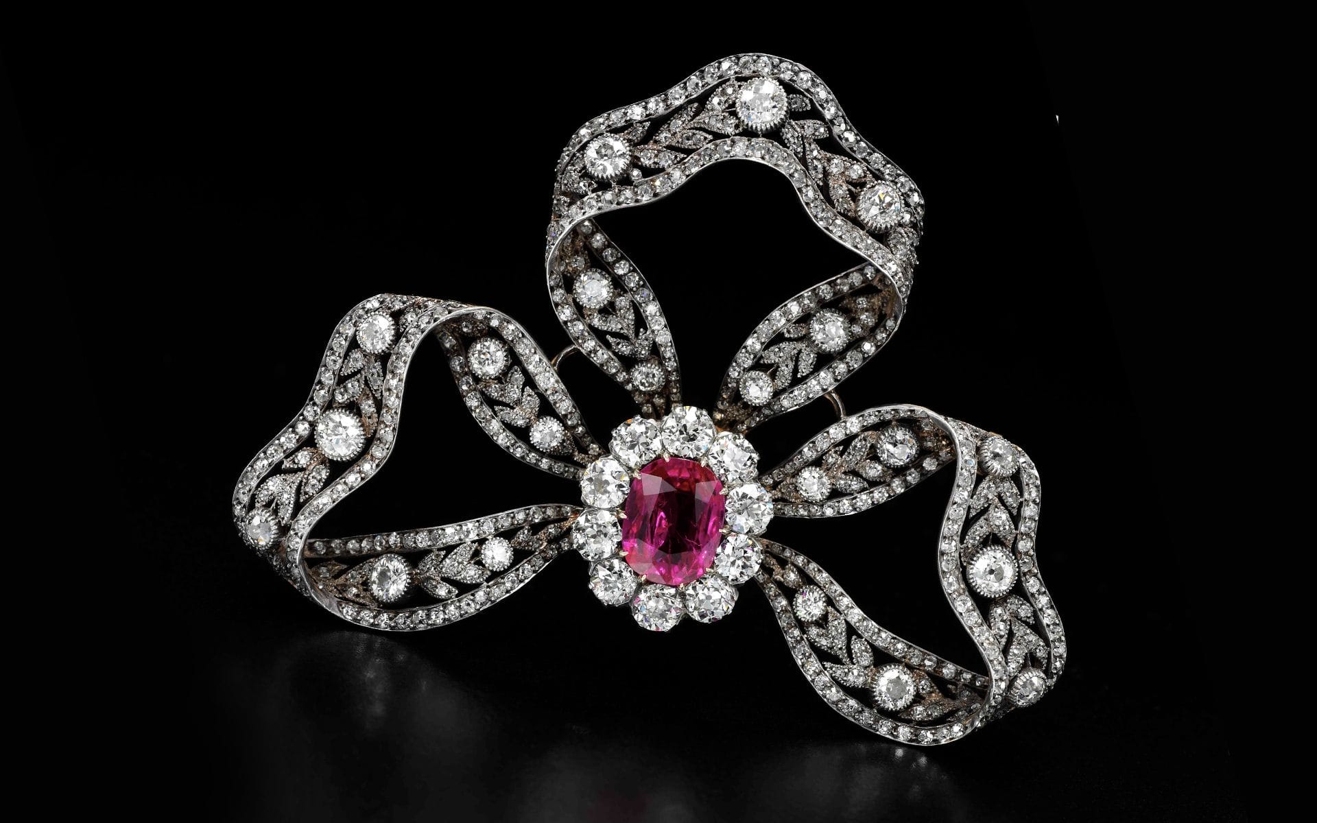 Elegant bow-shaped brooch with sparkling diamonds and a central vivid pink gemstone on a black background.