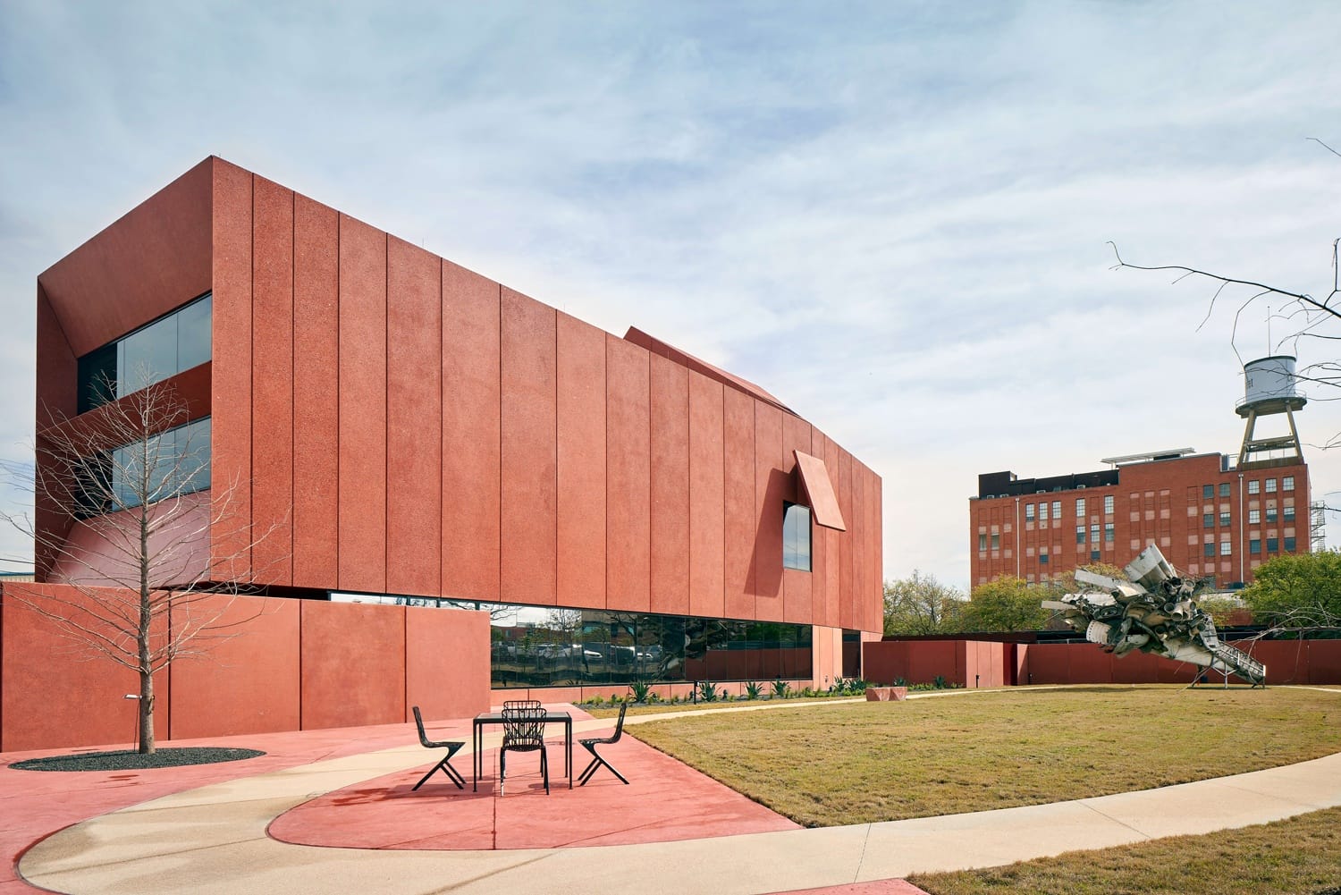 Modern red building with unique geometric design, large windows, and outdoor seating set against a clear sky and urban backdrop.