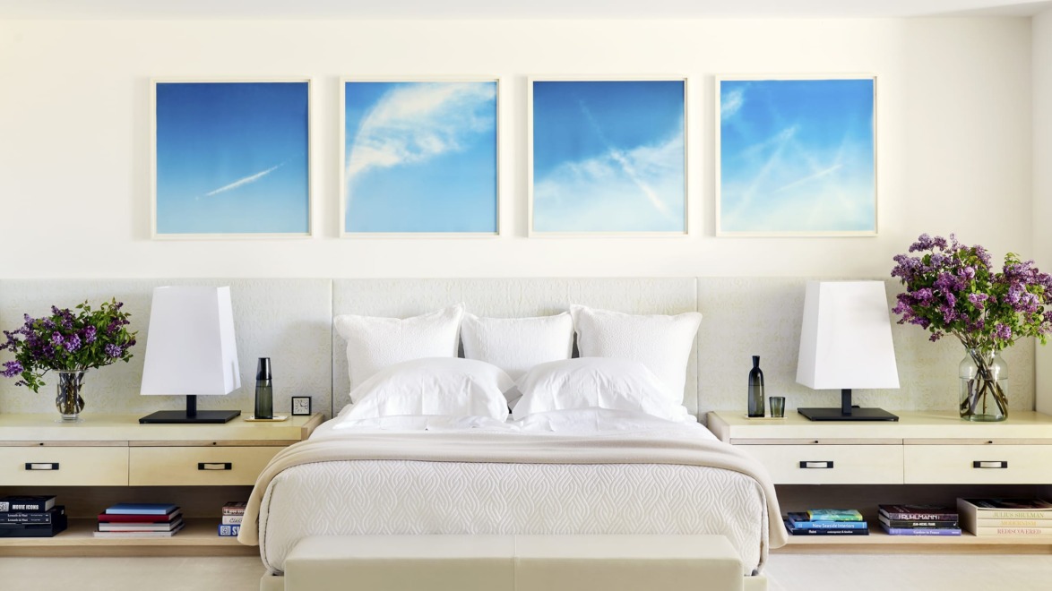Modern bedroom with white bedding, lavender flowers on nightstands, and four blue sky paintings on the wall above the bed.