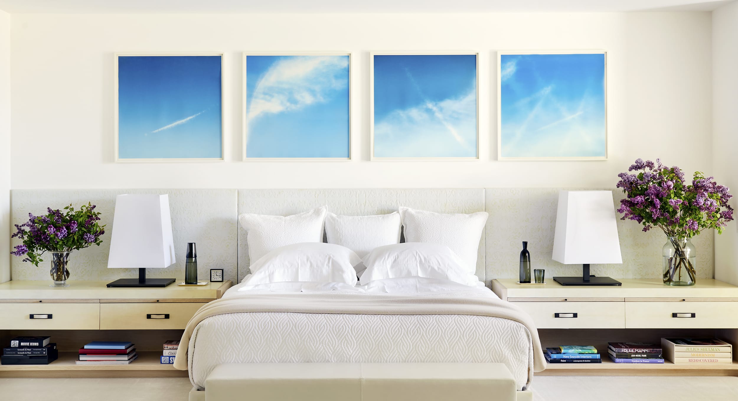Modern bedroom with white bedding, lavender flowers on nightstands, and four blue sky paintings on the wall above the bed.