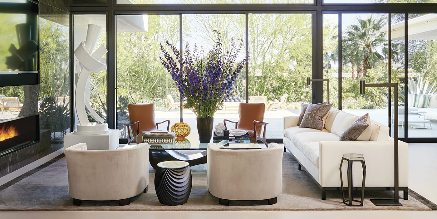 Modern living room with large windows, white sofas, brown chairs, a statue, and a view of greenery outside.