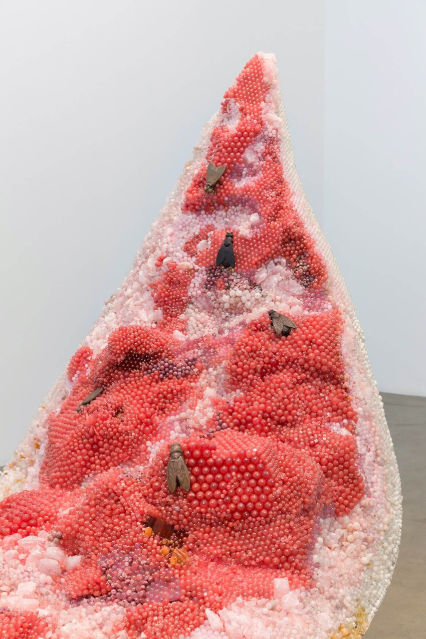 Sculpture featuring pink and red clustered spheres with scattered dark elements on a triangular form against a white background.