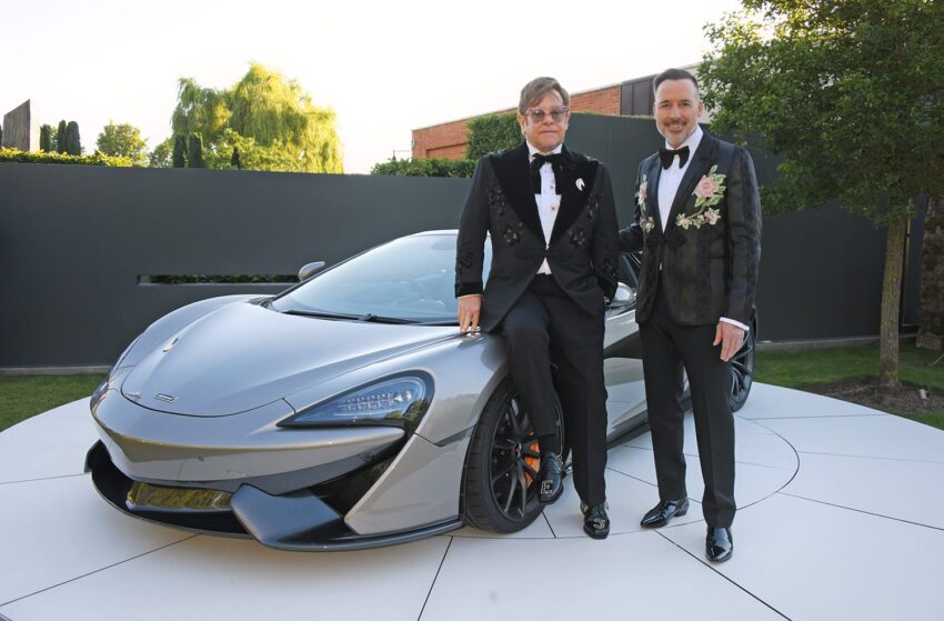 Two men in tuxedos standing in front of a sleek gray sports car outdoors.